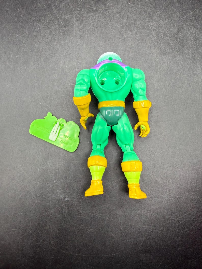 Op de afbeelding: Groene actiefiguur met gele accenten, waaronder handschoenen, laarzen en een riem. De figuur heeft een paarse helm en een groen accessoire. Vintage verzamelobject.