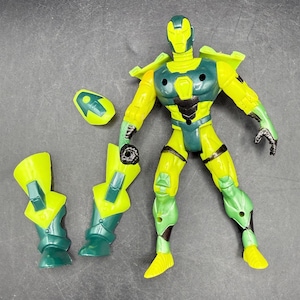 May include: A yellow and teal action figure with detachable parts. The figure has a helmet, chest armor, and leg armor. The figure is posed on a gray surface.