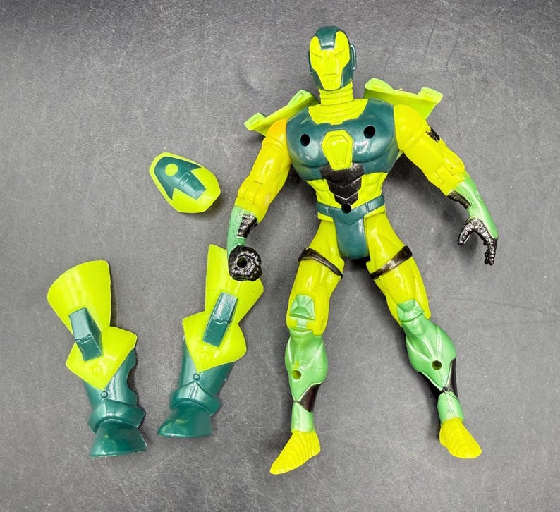 May include: A yellow and teal action figure with detachable parts. The figure has a helmet, chest armor, and leg armor. The figure is posed on a gray surface.
