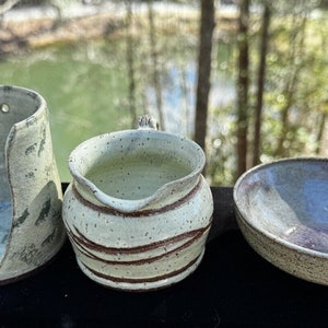 May include: Three handmade ceramic pieces in a light green and brown glaze. A tall, open-sided container, a small jug with a handle, and a shallow bowl.