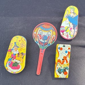 Peut inclure: Une collection de jouets en fer blanc vintage comprenant un bruiteur rond à visage de clown avec un manche rouge, une boîte en forme de goutte illustrée d'un artiste de cirque coloré et une boîte rectangulaire ornée de notes de musique.