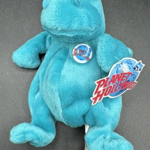 Puede incluir: Una rana de peluche azul turquesa con una etiqueta de Planet Hollywood adjunta. La rana tiene ojos grandes y una amplia sonrisa.