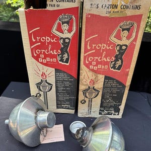 May include: Two vintage "Tropic Torches" patio product boxes with a retro design featuring a woman carrying fruit. Two silver metal torch fuel containers are in front of the boxes. The boxes are red and brown.