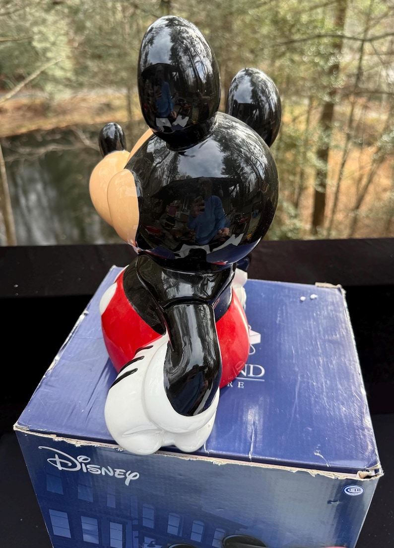 May include: A glossy black Mickey Mouse figurine with red shorts, white gloves, and tan ears. The figurine is on top of a blue Disney box. The figurine is a collectible item.