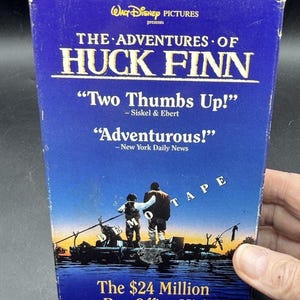 May include: A blue VHS tape box for The Adventures of Huck Finn by Walt Disney Pictures. The cover features two figures on a raft, the text Two Thumbs Up, Adventurous, and The $24 Million Box Office Hit, with the word DEMO TAPE printed diagonally.