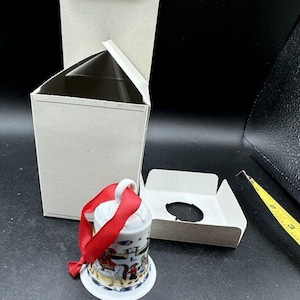 Könnte beinhalten: Eine weiße Porzellan-Glocke mit einem roten Band und einer bunten bemalten Szene von Menschen, die auf Pferden reiten. Die Glocke befindet sich in einer weißen Schachtel mit einem separaten weißen Einsatz.