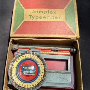 May include: A vintage Simplex Typewriter Number 100 in its original box. The typewriter is a small, portable model with a red and blue colour scheme. The box is made of cardboard and has a green and red design.