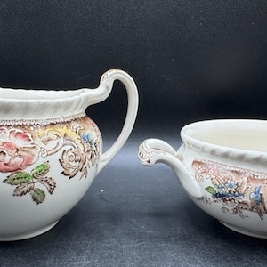 Op de afbeelding: Een crèmekleurige melk- en suikerpotset. De set heeft een bloemenontwerp met roze, bruine en blauwe accenten. De melkkan heeft een handvat en een tuit, terwijl de suikerpot twee handvatten heeft.