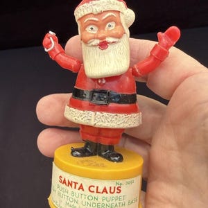 May include: Vintage Santa Claus push button puppet figurine. The red and white figure has a white beard, black belt, and is mounted on a yellow base. The base has text that reads "Santa Claus Push Button Puppet" and "Made in Hong Kong."