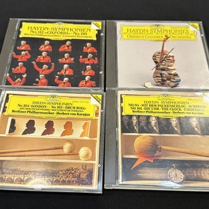 Puede incluir: Cuatro estuches de discos compactos con etiquetas amarillas y blancas. Las etiquetas muestran el texto "HAYDN SYMPHONIES" y enumeran los números de sinfonía y las orquestas. Los estuches están dispuestos en dos filas de dos.