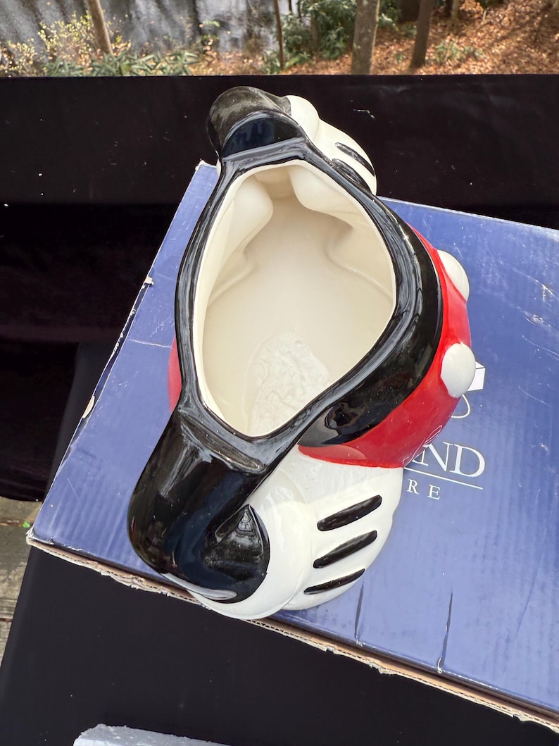 May include: A ceramic Mickey Mouse-shaped container. The container is primarily white, with a red body and black accents. The interior is white. The container is sitting on a blue box.
