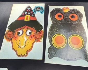 May include: Two vintage Halloween paper cutouts. One is a witch's face with a black hat, orange hair, and a long nose. The other is a black owl with orange eyes and a yellow and orange circle on its chest. A blue spider is next to the witch.