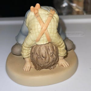 May include: A porcelain figurine of a child with brown hair, wearing a blue and yellow outfit with orange braces, bending over with their head down. The figurine is on a tan base.