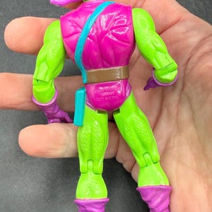 May include: A vintage action figure of the Green Goblin, a comic book villain. The figure is primarily green and purple, with a pink torso and a purple mask. The figure has a brown belt and a teal pouch. The figure is held in a hand.