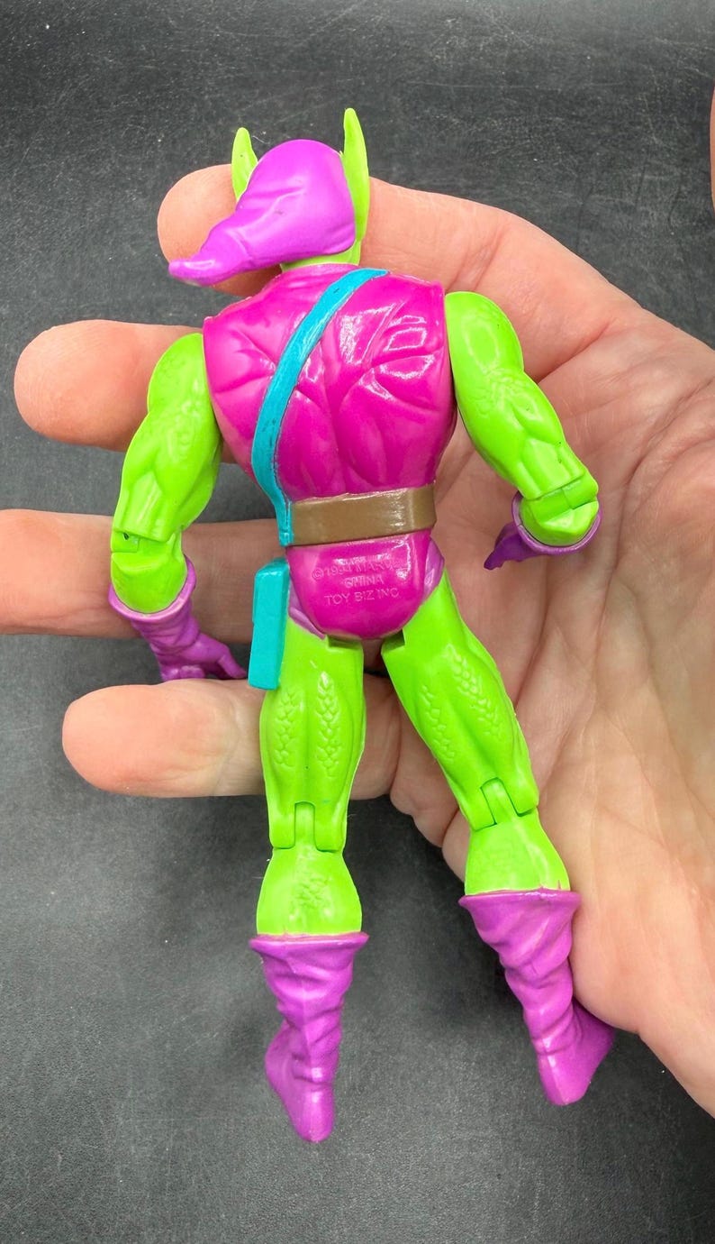 May include: A vintage action figure of the Green Goblin, a comic book villain. The figure is primarily green and purple, with a pink torso and a purple mask. The figure has a brown belt and a teal pouch. The figure is held in a hand.