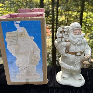 Puede incluir: Figura de cerámica de Papá Noel de color crema, sosteniendo una linterna y un saco de regalos. La figura está junto a su caja original, que presenta una imagen similar de Papá Noel. La caja tiene texto y una firma. Una decoración navideña festiva.