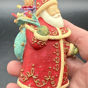 May include: A Santa Claus figurine wearing a red coat with gold scrollwork and cream-colored trim. Santa carries a teal sack filled with gifts and a small teddy bear. The figure has a white beard, a red hat with a cream pom-pom, and dark boots.