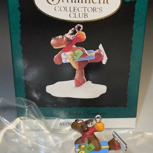 Peut inclure: Une figurine de renne brun portant une écharpe rouge et tenant une boîte cadeau bleue avec une étiquette verte indiquant "95". Le renne patine sur glace sur une base blanche avec un ruban bleu.