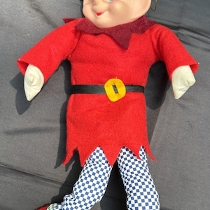 May include: A festive elf doll with a smiling face. The doll is dressed in a red felt tunic with a black belt and a yellow buckle. Its legs are patterned with a blue and white checkerboard design. The elf has a green and white chequered hat with a white pom-pom.