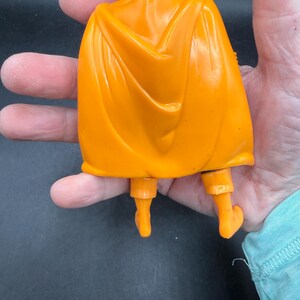 May include: Orange plastic action figure with a cape, boots, and a headpiece. The figure is a solid orange color and appears to be a superhero or fantasy character. The cape is draped over the figure's back.
