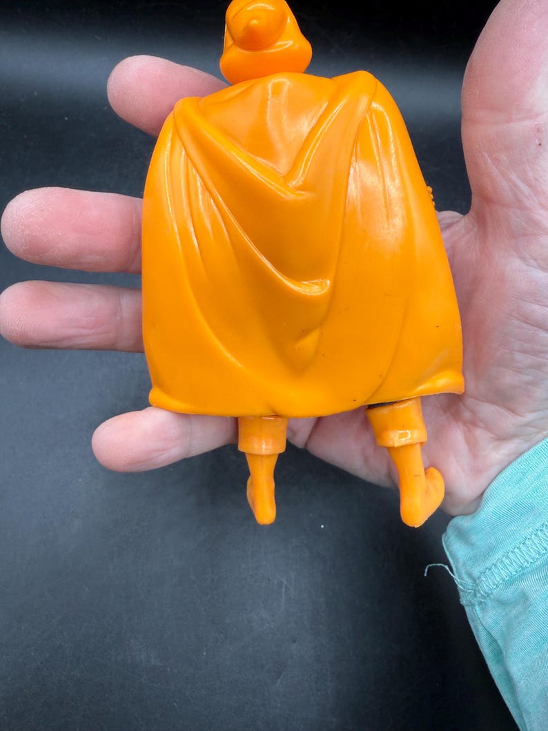 May include: Orange plastic action figure with a cape, boots, and a headpiece. The figure is a solid orange color and appears to be a superhero or fantasy character. The cape is draped over the figure's back.