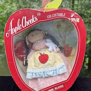 May include: A vintage "Apple Cheeks" doll in a red apple-shaped box. The doll wears a floral dress and a yellow apron with a red apple. The box reads "COLLECTABLE" and "MISS DEBBIE DELICIOUS".