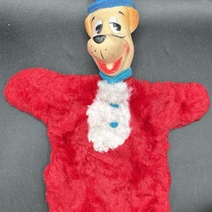 May include: A vintage hand puppet featuring a dog character with a blue top hat. The puppet has a red plush body with a white ruffled collar and blue accents. The dog's face is tan with a smiling red mouth.