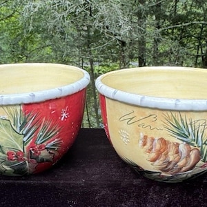 May include: Two ceramic bowls with a cream interior and a decorative exterior. The bowls have a red and yellow background with painted pinecones, holly, and the word "Warm". The rims are white.