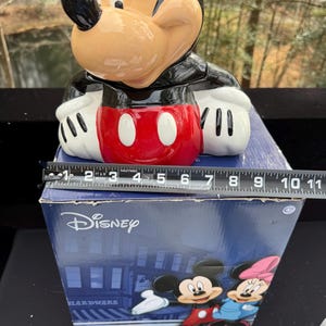 May include: A ceramic Mickey Mouse cookie jar with a black, red, white, and tan color scheme. The jar is sitting on a blue box with the Disney logo and a graphic of Mickey and Minnie Mouse. The jar measures approximately 10 inches in height.