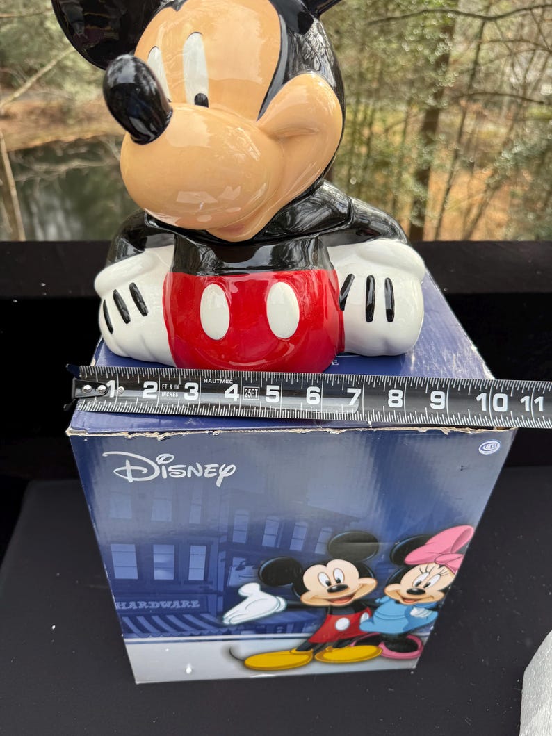 May include: A ceramic Mickey Mouse cookie jar with a black, red, white, and tan color scheme. The jar is sitting on a blue box with the Disney logo and a graphic of Mickey and Minnie Mouse. The jar measures approximately 10 inches in height.
