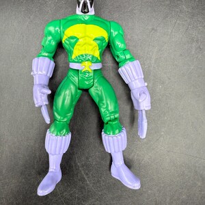 May include: A green action figure with a yellow chest emblem and a white and black mask. The figure has purple gloves, boots, and wrist cuffs. The toy is standing on a dark surface.