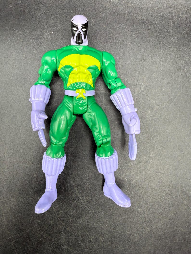 May include: A green action figure with a yellow chest emblem and a white and black mask. The figure has purple gloves, boots, and wrist cuffs. The toy is standing on a dark surface.