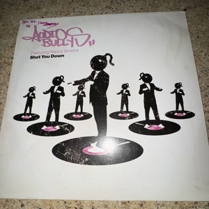 May include: A white vinyl record with a black and white illustration of a group of men in suits standing on record players. The text "Audio Bullys" is written in black graffiti-style font. The text "Featuring Nancy Sinatra" and "Shot You Down" are written in black font.