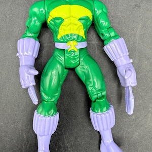 May include: A green action figure with a yellow emblem on the chest and a white and black mask. The figure has purple gloves, boots, and a belt. The toy is a collectible item.