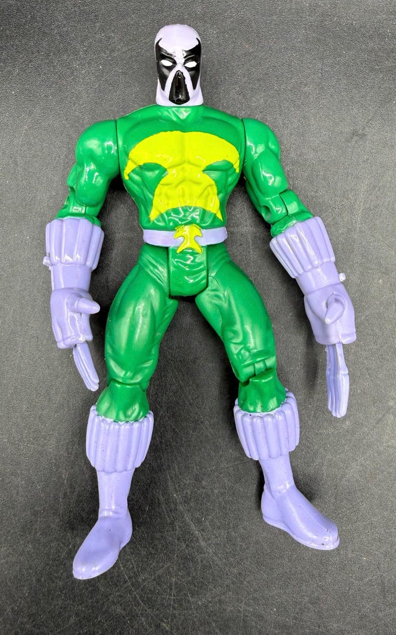 May include: A green action figure with a yellow emblem on the chest and a white and black mask. The figure has purple gloves, boots, and a belt. The toy is a collectible item.