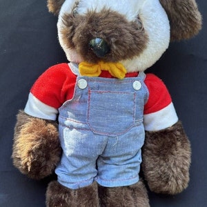 Op de afbeelding: Een bruine en witte pluche teddybeer met een rood shirt, een blauwe overall en een gele strik.