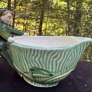 Puede incluir: Una jarra de crema de cerámica verde con una figura de elfo caprichosa que sostiene el asa. La jarra tiene un interior blanco y un exterior a rayas verde y blanco.