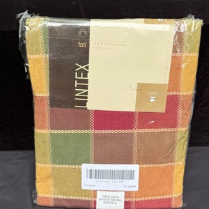 Może przedstawiać: Prostokątny obrus marki Lintex o wymiarach 152x304 cm wykonany w 100% z bawełny, zapakowany w przezroczystą folię. Tkanina posiada wielokolorowy wzór w kratę w odcieniach czerwieni, pomarańczu, żółci, brązu i zieleni, z dołączoną papierową etykietą.
