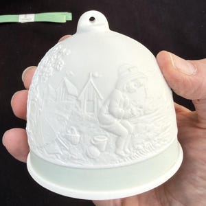 May include: White porcelain bell with a detailed relief scene of a person fishing near small huts and a basket. The bell has a light green base and a hole at the top for hanging. A decorative item.