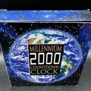 Può includere: Scatola con un design raffigurante la Terra blu e verde su uno sfondo stellato. Il testo "MILLENNIUM 2000 COUNTDOWN CLOCK" è visualizzato in bianco e oro, suggerendo un prodotto legato all'anno 2000.