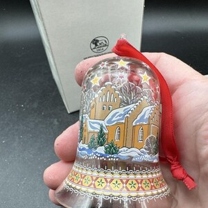 Könnte beinhalten: Klarer Glasglocken-Ornament mit rotem Band. Die Glocke zeigt eine Winterszene mit einer Kirche und Schnee. Der Boden der Glocke hat ein dekoratives Muster in Gelb, Rot und Weiß. Das Ornament befindet sich in einer weißen Box.