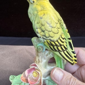 May include: A ceramic figurine of a yellow and green parakeet perched on a branch with a flower. The bird has black markings on its wings and head. The flower is pink and orange with green leaves.