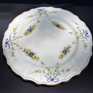 Könnte beinhalten: Ein runder weißer Keramikt weißer Keramikteller mit gewelltem Rand, verziert mit einem handgemalten Muster aus blauen Schleifen und Girlanden aus kleinen gelben und blauen Blumen, die in einer symmetrischen Rautenform um die Tellermitte angeordnet sind.