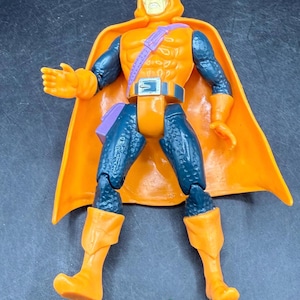 May include: Orange and blue action figure of a character with a hooded cowl and cape. The figure has orange boots, gloves, and a belt. A small orange pumpkin is at the figure's feet. The figure is a collectible toy.