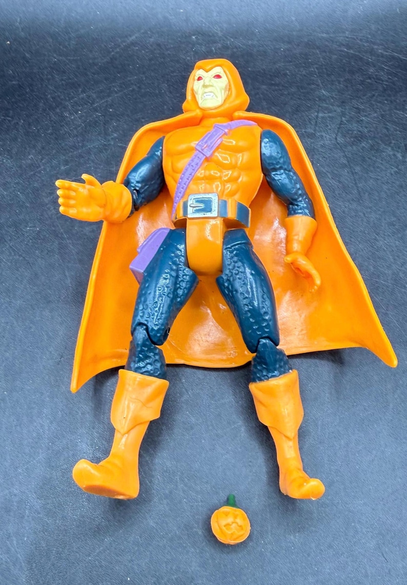 May include: Orange and blue action figure of a character with a hooded cowl and cape. The figure has orange boots, gloves, and a belt. A small orange pumpkin is at the figure's feet. The figure is a collectible toy.