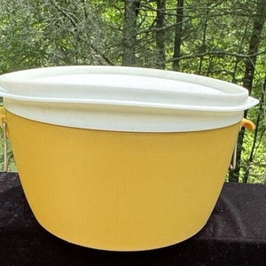 Op de afbeelding: Een gele plastic opbergcontainer met een wit deksel en twee handvatten. De container is rond en heeft een glad oppervlak.
