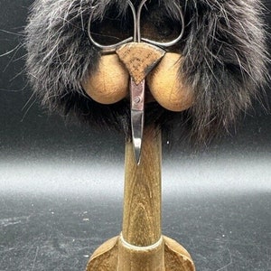 May include: A quirky wooden sculpture depicting a face with a fur 'hairdo', wooden 'cheeks', and a metal scissor-like element for a 'nose'. The sculpture is mounted on a wooden base, creating a unique decorative piece.