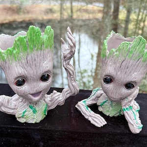 May include: Two Groot-style planters. The figures are light brown with a textured finish and green detailing on the head and body. They have large, black eyes. One has raised arms, the other rests its head on its hand.