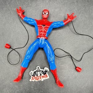 Puede incluir: Figura de acción de Spider-Man con su traje rojo y azul clásico, con cuerdas negras que simulan telarañas y extremos rojos. La figura está posando con los brazos extendidos. Una pegatina de Spider-Man es visible.