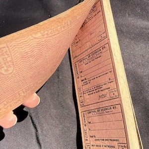 Peut inclure: Un livret de billets de la Central of Georgia Railway d'époque, de couleur brun rougeâtre. Les billets sont imprimés avec du texte et des détails pour un aller-retour, y compris l'itinéraire et la date. Le livret est ouvert, révélant plusieurs pages.